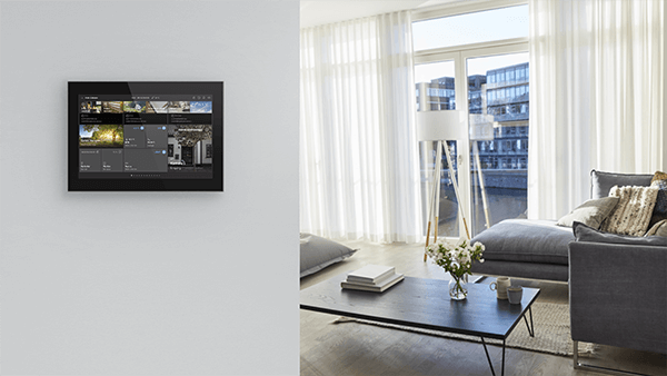 Das Bild zeigt ein Wohnzimmer mit einem Sofa, einem Couchtisch, einer Vase mit Blumen und großen Fenstern. An einer weißen Wand ist ein schwarzes Busch-OneTouch 7” angebracht.