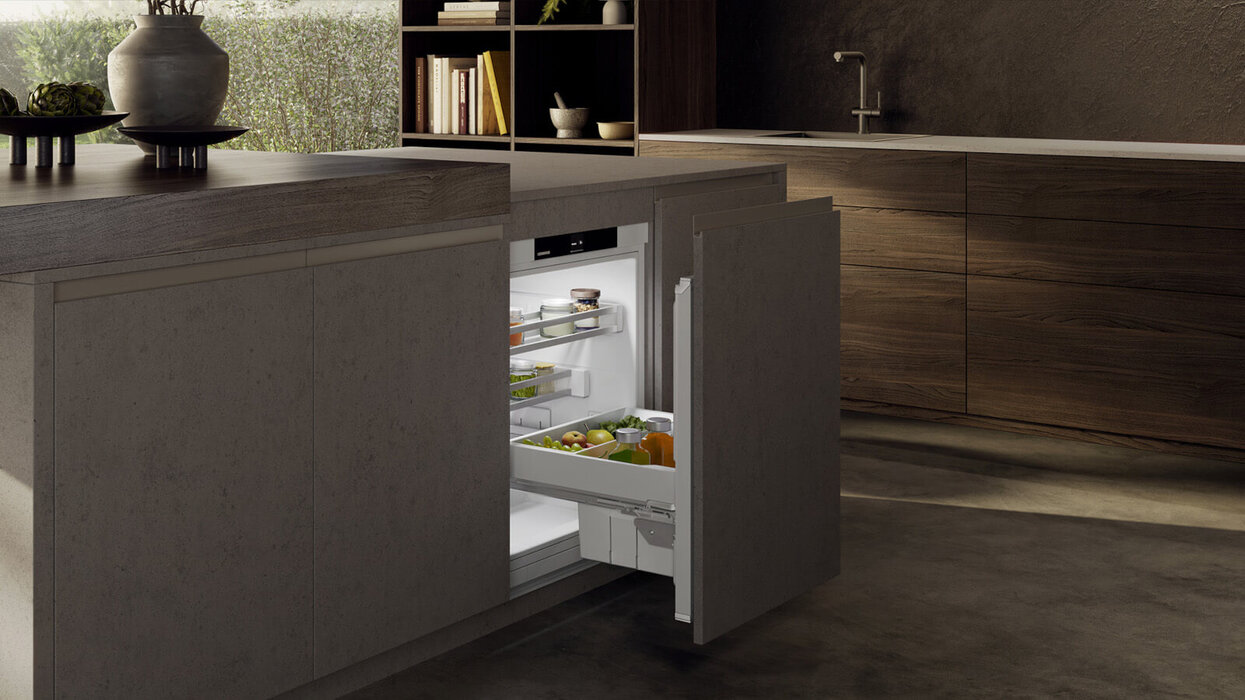 Moderne Küche mit großer Kochinsel, dunklen Holzmöbeln und raumhohen Fensterflächen. In die Insel ist ein geöffneter Unterbau-Kühlschrank integriert.