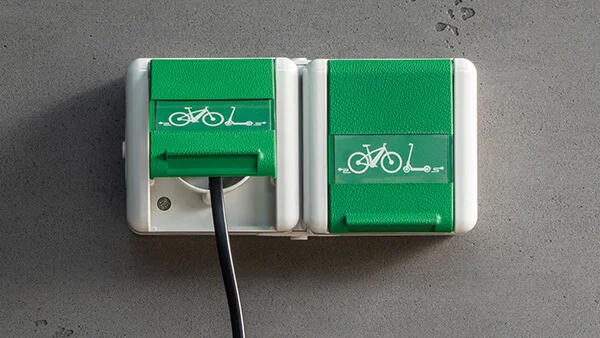 Zwei Steckdosen an der Wand montiert. Steckdose mit einer Klappe in der Farbe Grün und einem Fahrradsymbol in der Farbe Weiß.