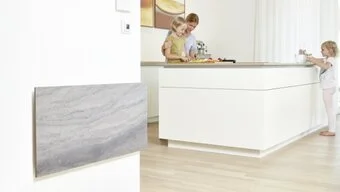 Zwei Personen stehen an einem Küchenblock. Im Vordergrund ist eine Infrarotheizung an einer Wand befestigt.