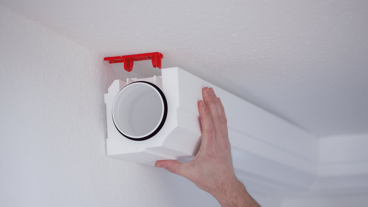 Eine Hand hält ein Lüftungsrohr mit Dämmung und roter Halterung an die Zimmerdecke.
