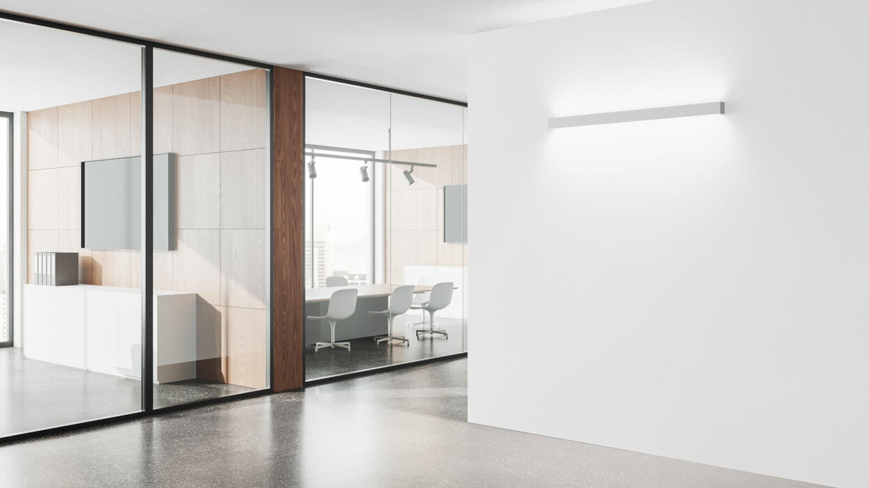 Linientreu – Sylvania Otao Linear Ein heller Büroraum mit Glaswänden und einem Konferenztisch im Hintergrund. An einer weißen Wand im Vordergrund ist eine schlanke Wandleuchte montiert, die Licht nach oben und unten abstrahlt.