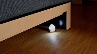 Das Bild zeigt ein kleines Nachtlicht mit Bewegungsmelder, das unter einem Bett auf einem Holzboden leuchtet.