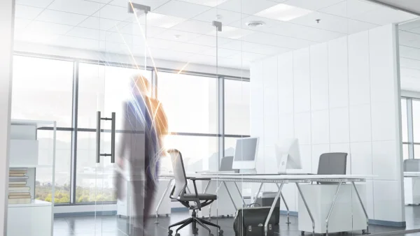 Das Bild zeigt einen modernen Büroraum mit Glaswand und Glastür. Die Seitenwand zum Nachbarbüro ist wie die Büroausstattung ist weiß. Eine Person mit Aktentasche hat den Raum durch die offene Glastür betreten. Die Person ist unscharf dargestellt. Die Person wird auf der linken Seite mit blau-weißen und auf der rechten Seite von gelb-roten Lichtstreifen beleuchtet.