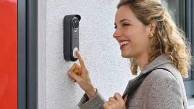 Eine Person betätigt die Klingeltaste einer modernen Video-Türsprechanlage mit integrierter Kamera, die an einer Hauswand neben einer Eingangstür montiert ist.