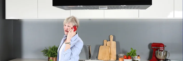 Das Bild zeigt eine ältere Person, die in einer modernen Küche telefoniert. Im Hintergrund sind Küchenkräuter und Kochutensilien zu sehen.