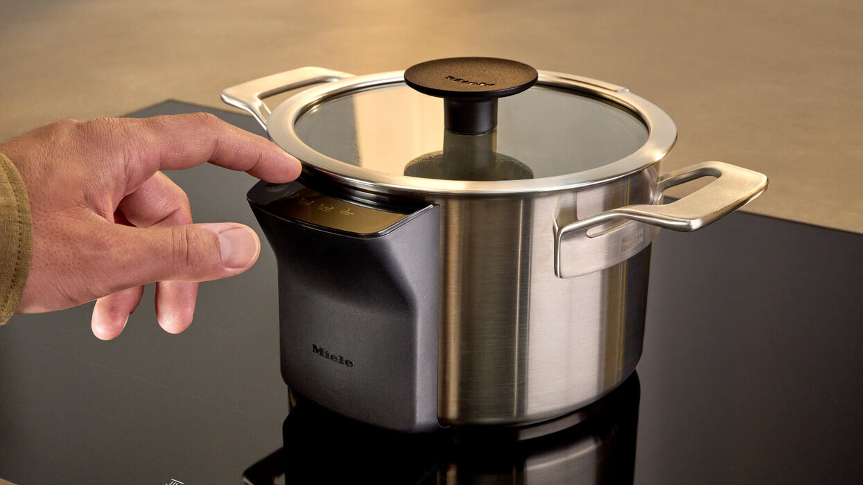 Das Bild zeigt einen modernen Kochtopf auf einem Induktionskochfeld, bei dem eine Hand das Bedienfeld am Topf berührt.