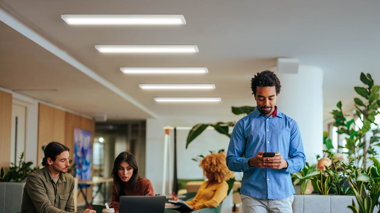 Ein moderner Büroraum mit mehreren Menschen: Zwei sitzen am Laptop und arbeiten zusammen, während ein Mann im Vordergrund auf sein Smartphone schaut. Pflanzen und helle Beleuchtung sorgen für eine freundliche Atmosphäre.