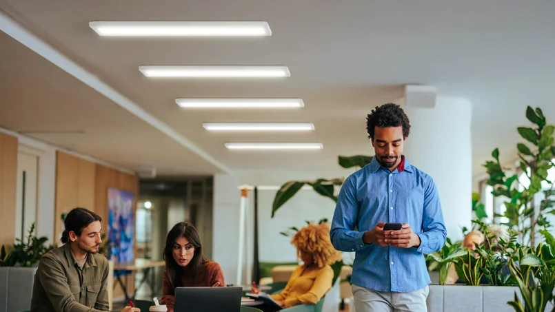 Ein moderner Büroraum mit mehreren Menschen: Zwei sitzen am Laptop und arbeiten zusammen, während ein Mann im Vordergrund auf sein Smartphone schaut. Pflanzen und helle Beleuchtung sorgen für eine freundliche Atmosphäre.