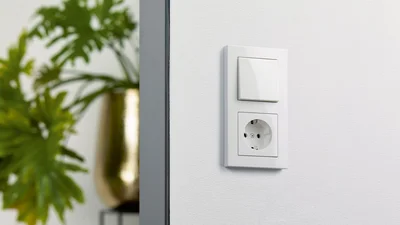 An einer weißen Wand ist eine weiße Kombination aus Lichtschalter und Steckdose angebracht. Im unscharfen Hintergrund links ist eine grüne Pflanze in einem goldenen Topf zu sehen, die durch einen dunklen Türrahmen oder eine Säule getrennt ist.