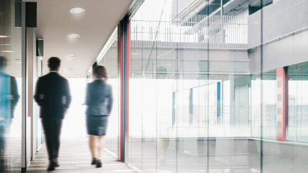 Zwei Personen gehen nebeneinander einen hellen Gang entlang. Die Wände rechts und links des Ganges sind aus Glas. Im Glas auf der rechten Seite spiegelt sich die Umgebung. Die Decke des Ganges wird durch Downlights beleuchtet.