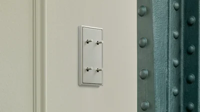 Das Bild zeigt einen eleganten Lichtschalter mit vier Kippschaltern in Metalloptik an einer hellen Wand, neben einem Stahlträger mit Nieten.