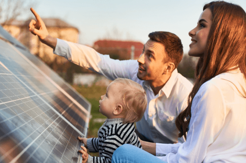 Das Bild zeigt eine Frau, einen Mann und ein Kleinkind, die gemeinsam im Freien Solarmodule betrachten. Der Mann in der Mitte deutet mit dem Finger nach oben auf die Paneele, während die Frau rechts und das Kleinkind links von ihm stehen.