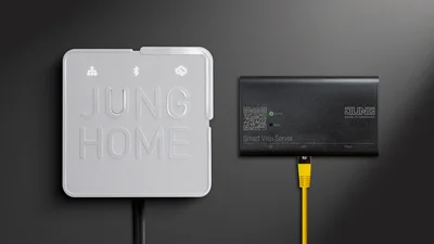 Nahaufnahme von zwei Geräten an einer Wand: das JUNG HOME Gateway (weiß) und der Smart Visu Server (schwarz) mit angeschlossenem gelbem Netzwerkkabel.