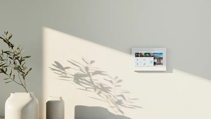 Das Bild zeigt einen weißen Busch-OneTouch 7" an einer hellen Wand. Daneben steht ein Sideboard aus hellem Holz, auf dem zwei weiße Vasen stehen. In einer Vase steckt ein Zweig. 