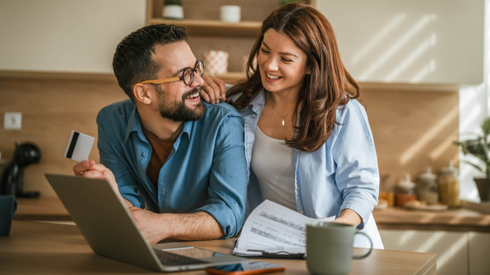 Zwei Personen stehen in einer Küche an einem Tisch mit Laptop, Unterlagen und einer Tasse. Eine Person hält eine Karte in der Hand, während beide auf den Bildschirm blicken.